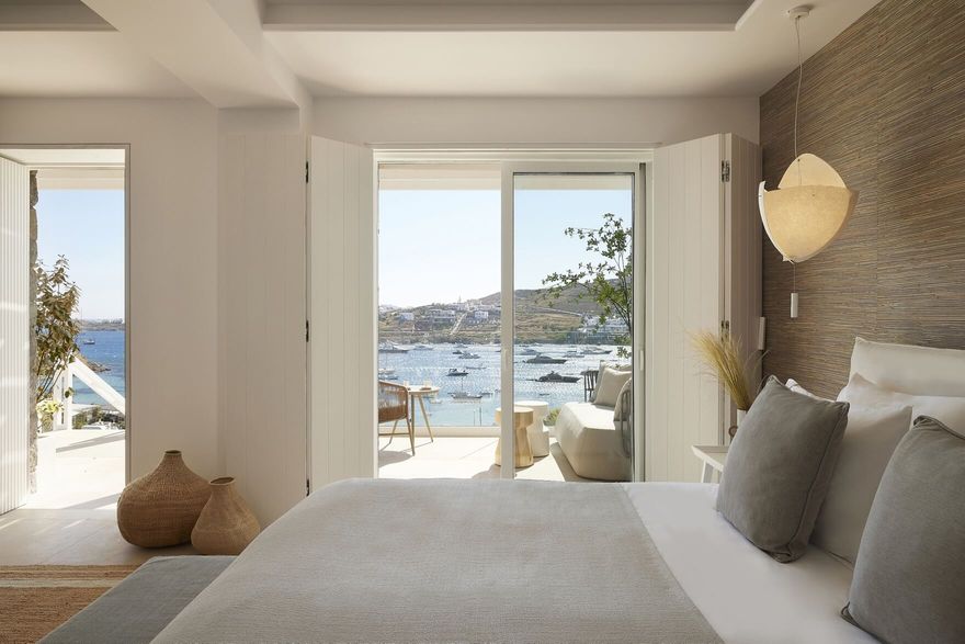 Luxury suite at Once In Mykonos hotel with soft neutral decor, opening onto a private terrace with panoramic views of Ornos Bay and anchored yachts