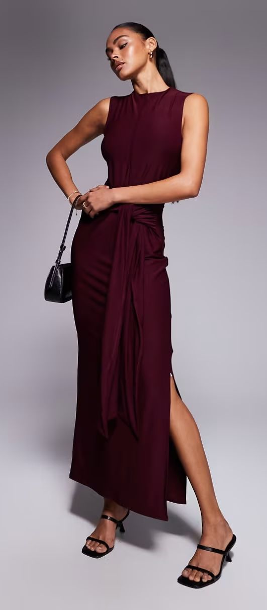 Model in a sleeveless burgundy maxi dress with draped waist detail and thigh split, holding a black bag