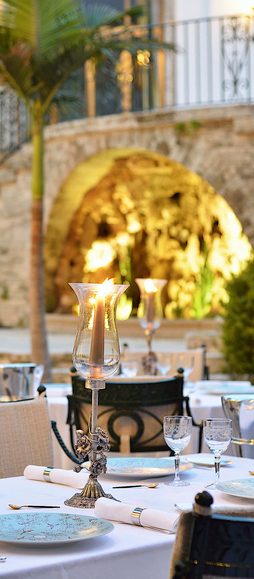 Outdoor terrace at Candlelight Restaurant at Hostal de La Gavina in Costa Brava, featuring elegantly dressed tables, candlelit dining, palm trees and historic stone architecture, creating a romantic setting for couples.