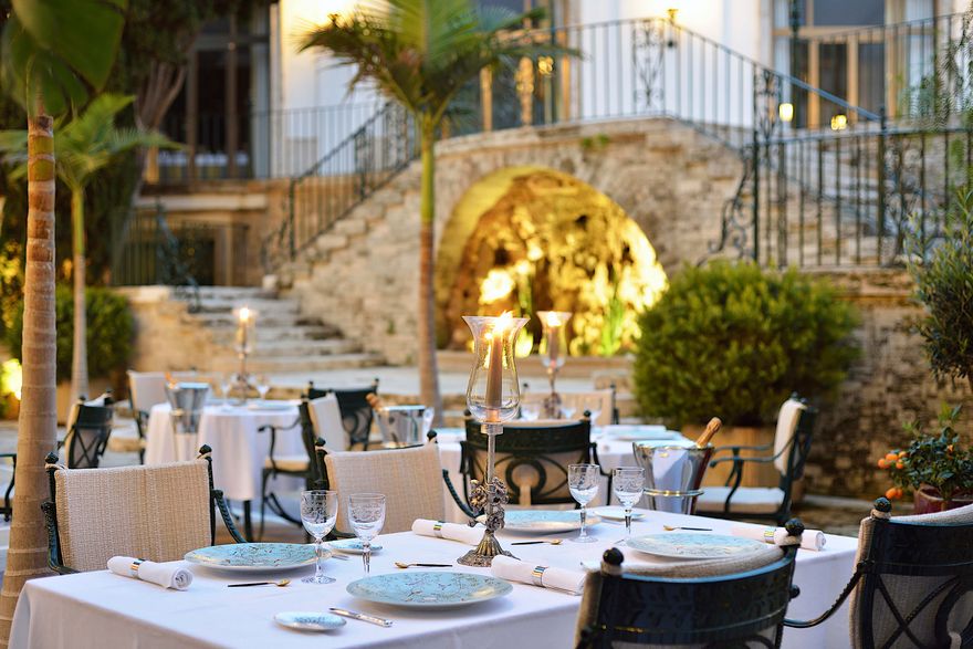 Outdoor terrace at Candlelight Restaurant at Hostal de La Gavina in Costa Brava, featuring elegantly dressed tables, candlelit dining, palm trees and historic stone architecture, creating a romantic setting for couples.