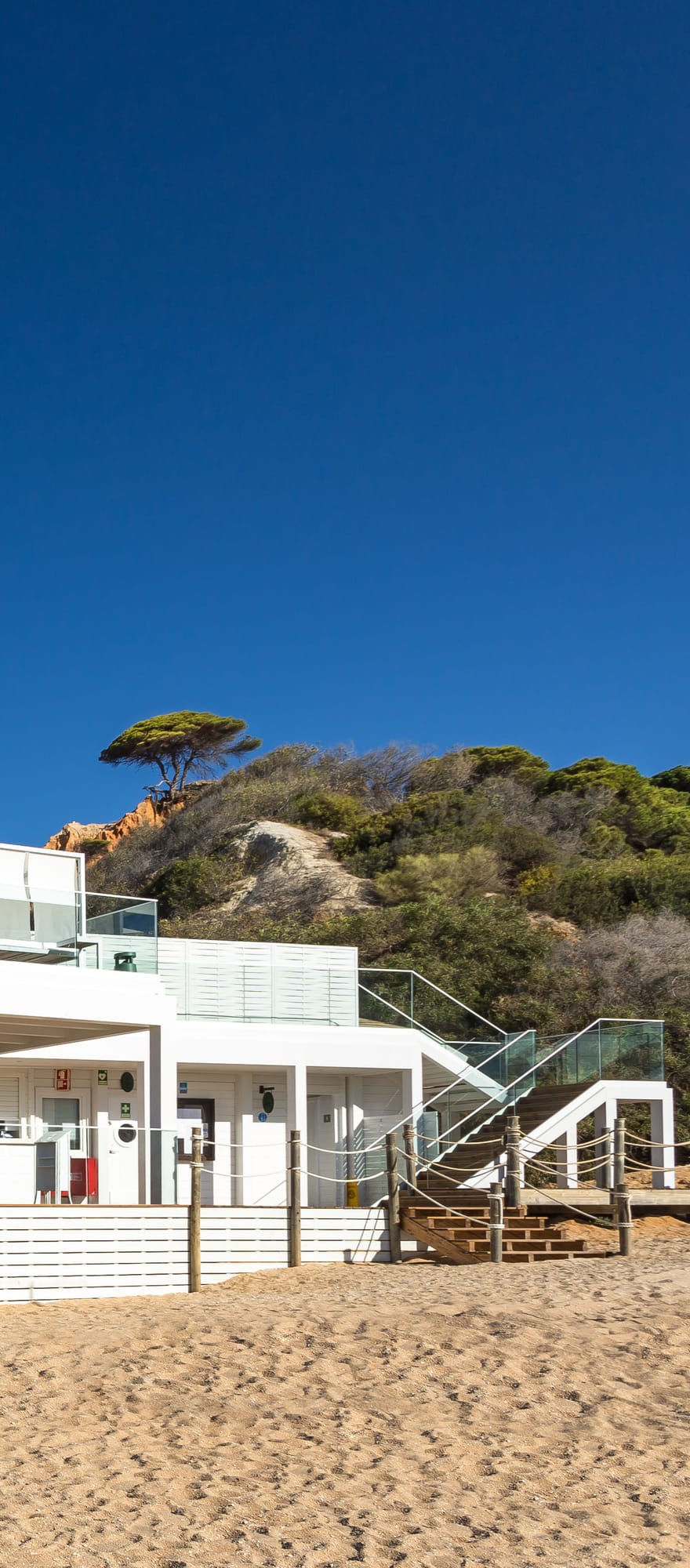 Maré beachfront restaurant at Pine Cliffs Resort in the Algarve, Portugal, featuring a white seaside terrace with parasols overlooking Praia da Falésia beach and the Atlantic Ocean.