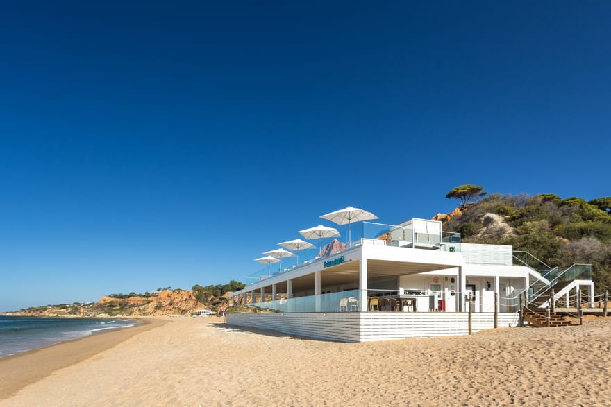 Maré beachfront restaurant at Pine Cliffs Resort in the Algarve, Portugal, featuring a white seaside terrace with parasols overlooking Praia da Falésia beach and the Atlantic Ocean.