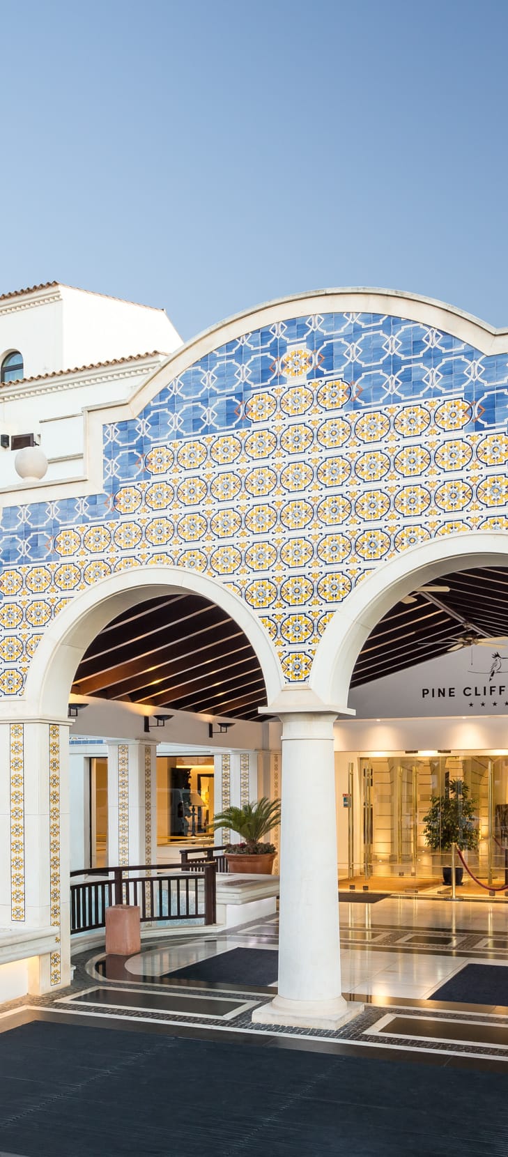 Main entrance of Pine Cliffs Hotel in the Algarve, Portugal, featuring traditional blue and yellow Portuguese tiles, grand white archways, palm trees and decorative water features.