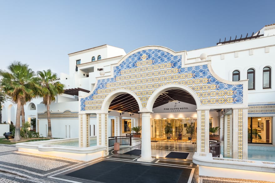 Main entrance of Pine Cliffs Hotel in the Algarve, Portugal, featuring traditional blue and yellow Portuguese tiles, grand white archways, palm trees and decorative water features.