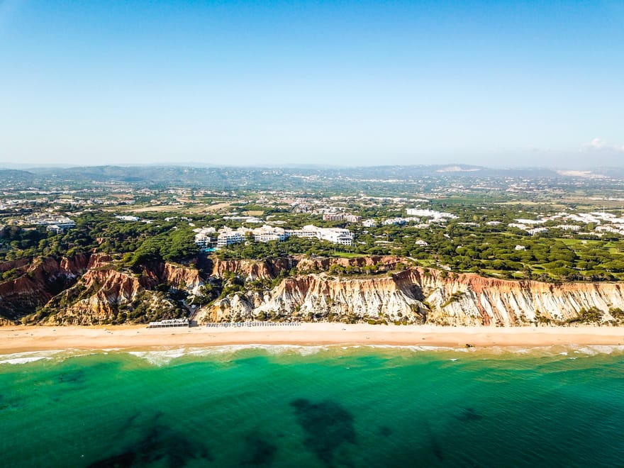 Aerial view of Pine Cliffs Resort overlooking Praia da Falésia beach in the Algarve, Portugal, featuring dramatic red and golden cliffs, turquoise Atlantic waters and a long stretch of sandy coastline.
