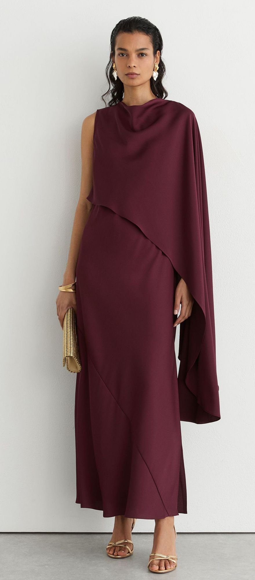 Woman in a burgundy one-shoulder cape maxi dress with gold clutch and sandals