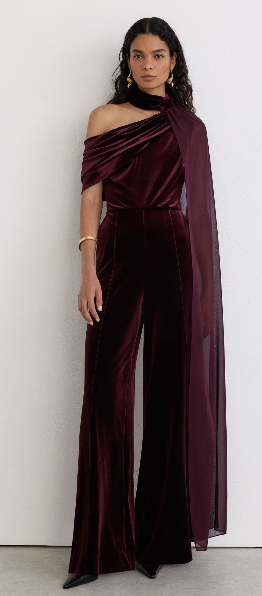 Woman in a burgundy velvet one-shoulder jumpsuit with flowing chiffon cape detail