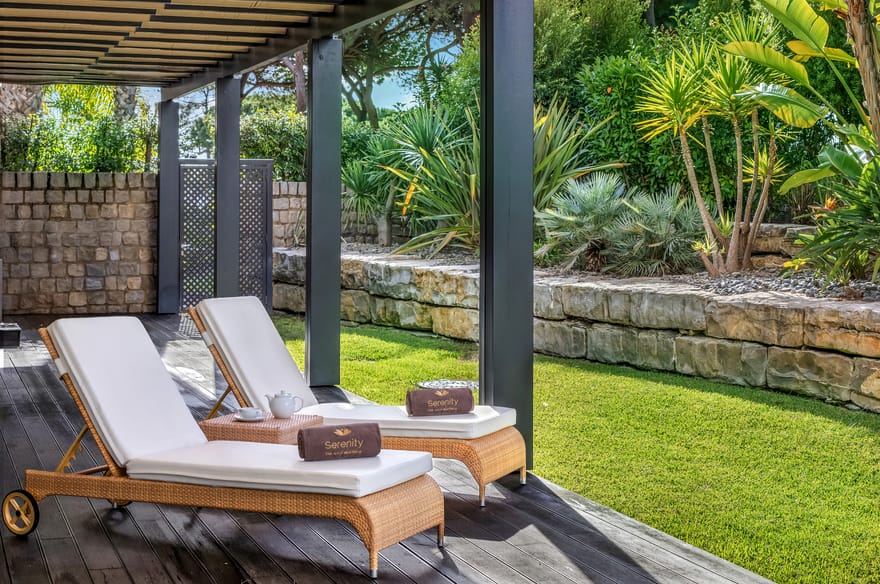 Outdoor relaxation area at Serenity Spa, Pine Cliffs Resort in the Algarve, Portugal, featuring two luxury sun loungers on a shaded wooden terrace surrounded by tropical gardens.